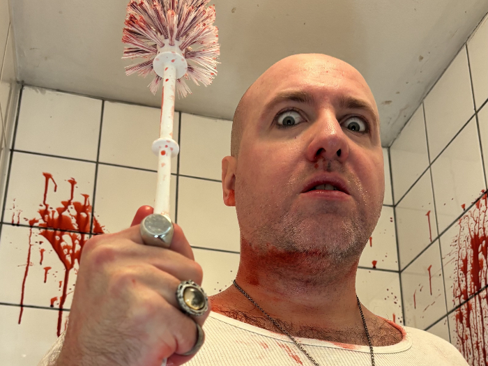 Schauspieler des König Ubu hält eine blutverschmierte Klobürste als Zepter